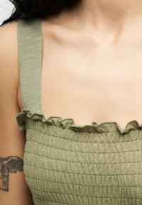 Γυναίκα που φοράει τοπ χωρίς μανίκια σε υφή ελιάς με βολάν στο ντεκολτέ και φαίνεται τατουάζ με πουλί στο πάνω μέρος του βραχίονα.