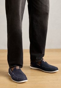 Jūriniai slip-on batai su tekstūruotu audinio viršumi, balta gumine padais ir maža odinė detalė ant kulno, dėvimi su juodais džinsais.