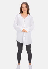 White button-up shirt made of textured fabric, featuring a collar, long sleeves, and front placket. Paired with black leggings and white sneakers.