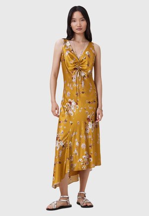 Abito di raso giallo con un motivo floreale, scollatura a V profonda, cordoncino regolabile in vita e orlo asimmetrico; la modella indossa sandali piatti.