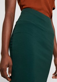Close-up van een persoon die een groene high-waisted rok en een roestkleurige geribbelde top draagt, met de handen en het midden van het lichaam zichtbaar.
