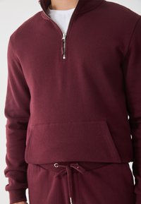 Burgundská mikina na zip z měkké tkaniny s klokanskou kapsou a ribbenovými manžetami. Má stříbrný zip a volný střih.