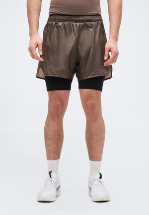 Άνδρας που φοράει καφέ άνετα σορτς πάνω από μαύρα συμπιεστικά σορτς, λευκές κάλτσες και λευκά αθλητικά παπούτσια, στέκεται με τα χέρια χαλαρά.