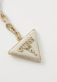 Baltas trikampis odinis raktų pakabukas su tekstūruota paviršiumi, auksinėmis detalėmis, įspaustu "GUESS USA" logotipu ir dekoratyvine klausimo ženklo detale.