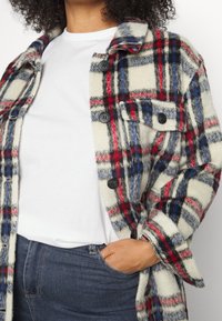 Ruudullinen ylimitoitettu takki vaaleassa, punaisessa ja sinisessä sävyssä. Valmistettu pehmeästä kankaasta, jossa on isot mustat napit ja kaksi etutaskua.