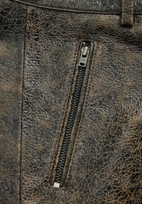 Gros plan sur une fermeture éclair noire verticale sur un tissu en cuir brun foncé usé et texturé, avec des coutures visibles autour de la fermeture.