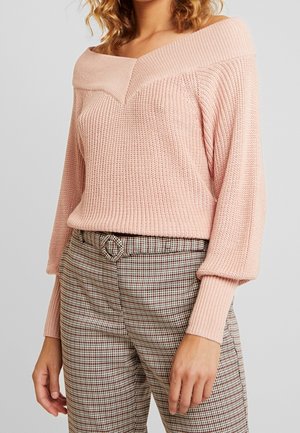 Frau trägt einen hellrosa schulterfreien Strickpullover mit Ballonärmeln und hoch taillierte karierte Hose mit passendem Gürtel.