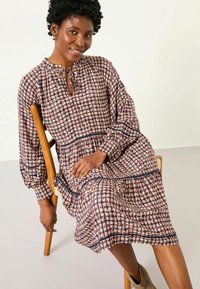 Gemustertes Midikleid aus weichem Stoff, mit geraffter Taille, langen Puffärmeln und einem Schlüssellochausschnitt mit Bindebändern. Dunkelblaue Akzente.