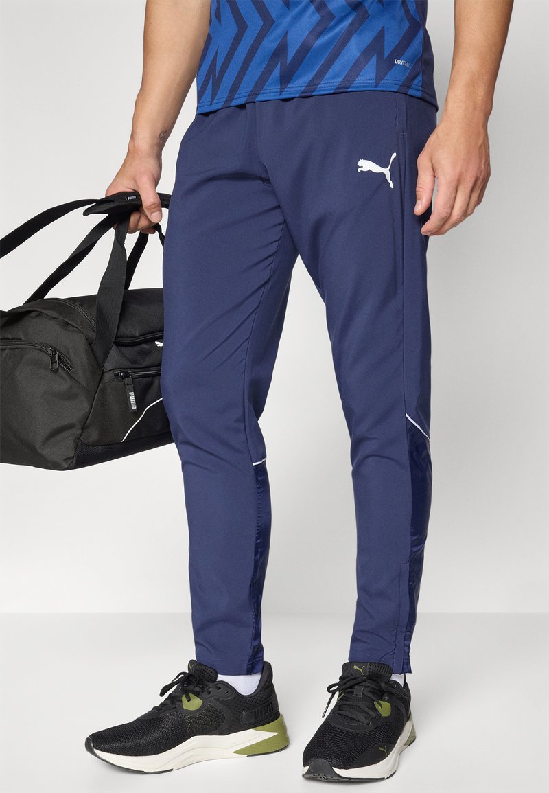 Pantalones deportivos ajustados de color navy, con paneles laterales contrastantes, acentos blancos y un logo de Puma. Lleva un bolso de viaje negro y zapatillas negras.