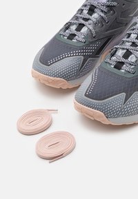 Šedé sportovní boty s texturovaným svrškem a světle růžovými podrážkami, doplněné růžovými a fialovými šňůrkami s tečkovaným vzorem na bílém pozadí.