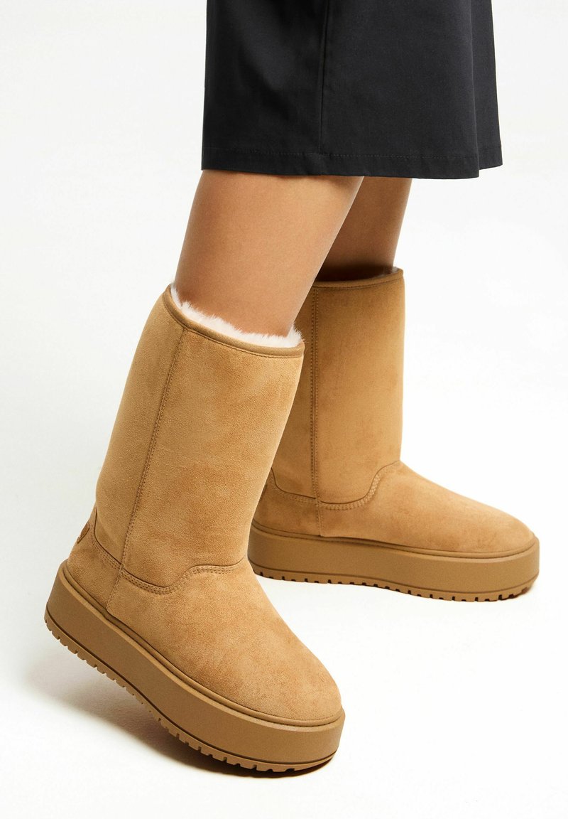 Tan suede ankelstøvler med en blød, hvid pelsforing øverst, der har en flad gummisål og en rund tådesign.