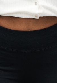 Vue de la taille d'une personne portant un haut blanc boutonné et un pantalon ou legging noir taille haute.