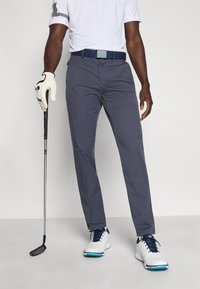 Pantalones de golf grises hechos de material ligero; cuentan con corte de pierna recta, bolsillos laterales y un cinturón con hebilla metálica. Combinados con zapatos blancos.