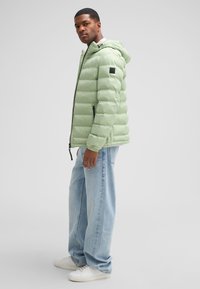 Giacca puffer verde chiaro con cappuccio, frontale con zip e patch logo nero. Indossata con jeans blu larghi e scarpe da ginnastica bianche.