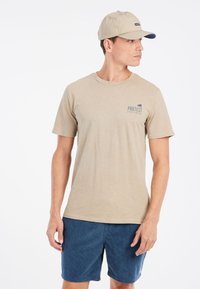 Camiseta beige de manga corta con cuello redondo y un pequeño logo azul "PROTEST". Combinada con pantalones cortos de pana azul y una gorra a juego.