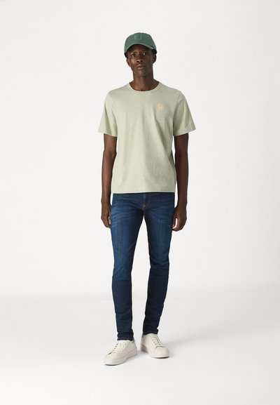 T-shirt vert clair à manches courtes avec un petit logo, associé à un jean slim bleu foncé et des baskets blanches. Une casquette verte assortie complète le look.