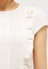 Světle růžová blůzka s krátkým rukávem, kulatým výstřihem a vertikálními volány podél přední části a okrajů rukávů. Hladká, strukturální látka.