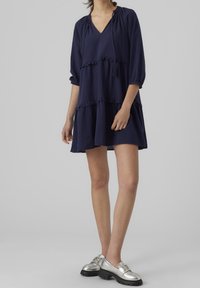 Kvinna klädd i en lös, rutig marinblå klänning och silverfärgade chunky loafers med dekorerade spännen, stående mot en enkel grå bakgrund.