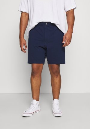 Homme portant un short bleu marine, des baskets blanches montantes et un t-shirt blanc ample, debout devant un fond clair uni.