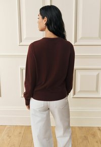 Bordo krāsas adīts džemperis arapaļo kakla izgriezumu un garām piedurknēm, kombinēts ar baltām plašām džinsa biksēm. Gluda tekstūra un pieguļoša vidukļa daļa.