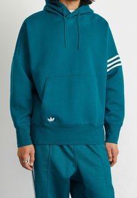 Teal bomullsblandning hoodie med en framficka, justerbar dragskohet och tre vita ränder på vänster ärm. Logotyp vid fållen.