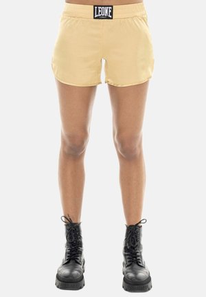 Person trägt beige Sportshorts mit dem Label "LEONE 1947" und schwarze Schnür-Kampfboots und steht vor einem einfarbigen hellen Hintergrund.