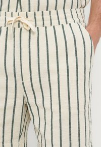 Gros plan sur un pantalon beige à rayures verticales noires, avec une taille à cordon et une main posée dans la poche droite.