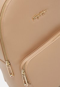 Beżowy skórzany plecak z złotymi zamkami i okuć. Posiada wyraźne logo marki w kolorze złotym. Gładka tekstura i minimalistyczny design.