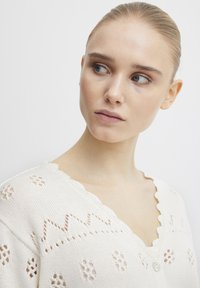 Cremefarbener Strickcardigan mit gewelltem Ausschnitt, Filigranmuster und einem einzelnen Knopfverschluss.