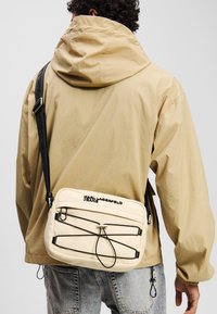 Helle beige Umhängetasche mit schwarzen Akzenten und Kordelzugdetail, ausgestattet mit einer glatten Textur und Logo. Über einer beigen Jacke getragen.
