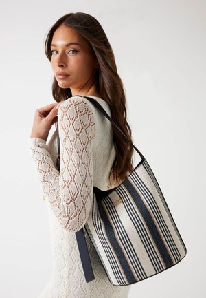 Gestreepte canvas emmer tas in beige, marineblauw en perzik, met leren accenten en een enkele schouderband. Soepele textuur met een gestructureerde vorm.