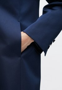 Tmavomodré sako s hladkou textúrou látky a mierne prispôsobeným strihom. Má viditeľnú vrecko a gombíkové detaily na manžete.