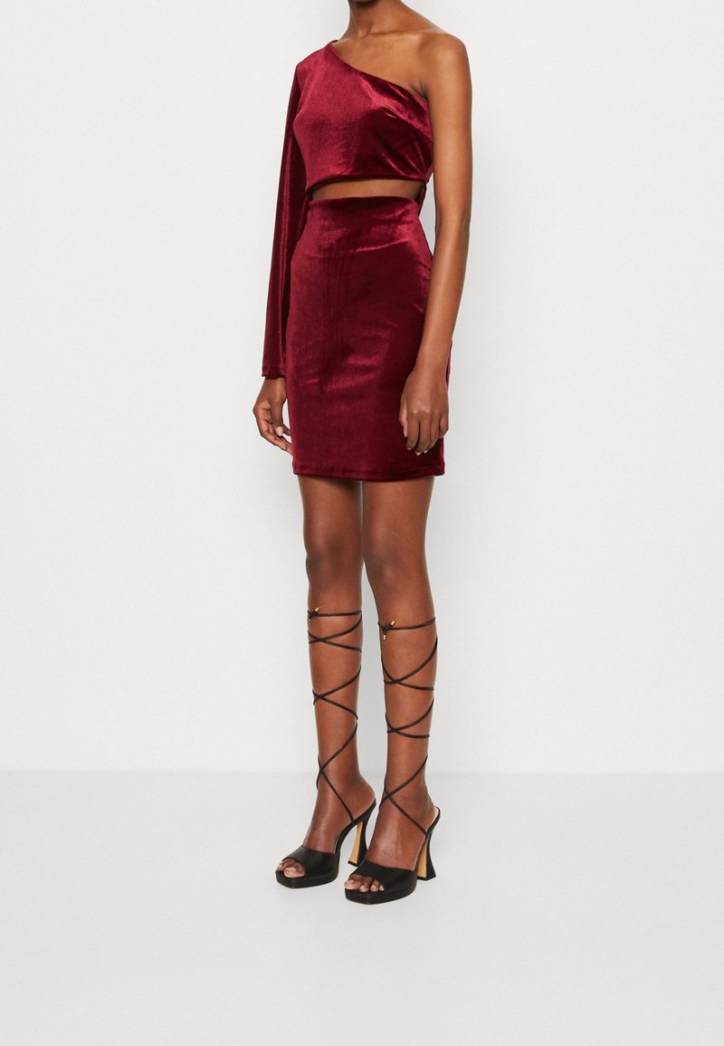 Ensemble deux-pièces en velours bordeaux comprenant un haut court asymétrique à une épaule et une minijupe ajustée, assorti à des sandales noires à talons hauts.