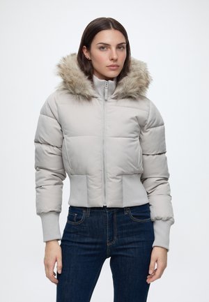Donna che indossa una giacca imbottita grigio chiaro con cappuccio bordato di pelliccia e jeans blu scuro, in piedi davanti a uno sfondo bianco semplice.