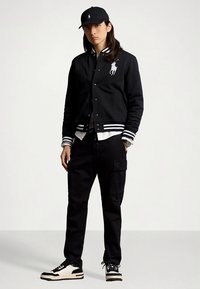 Czarna kurtka varsity z białymi paskami i haftowanym logo, noszona na białej koszuli. Dopełnieniem stylizacji są czarne spodnie typu cargo oraz czarno-białe sneakersy.