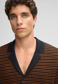 Polo shirt a righe in nero e arancione, con colletto nero, chiusura a bottoni e tessuto testurizzato dal fit rilassato.