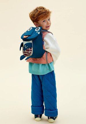 Red-haired child wearing layered clothes and a blue turtle-shaped backpack, looking back and smiling against a plain background.