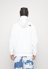 Felpa bianca con cappuccio regolabile, caratterizzata da un logo nero sulla spalla posteriore. Abbinata a jeans tie-dye blu e bianchi.