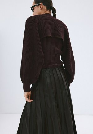 Femme portant un pull côtelé violet foncé avec des manches bouffantes et une longue jupe plissée noire, vue de dos sur un fond blanc.