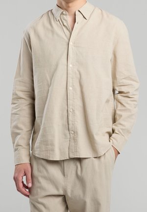 Mand iført beige langærmet skjorte med knapper, med den ene hånd i lommen mod en ensfarvet lys grå baggrund.