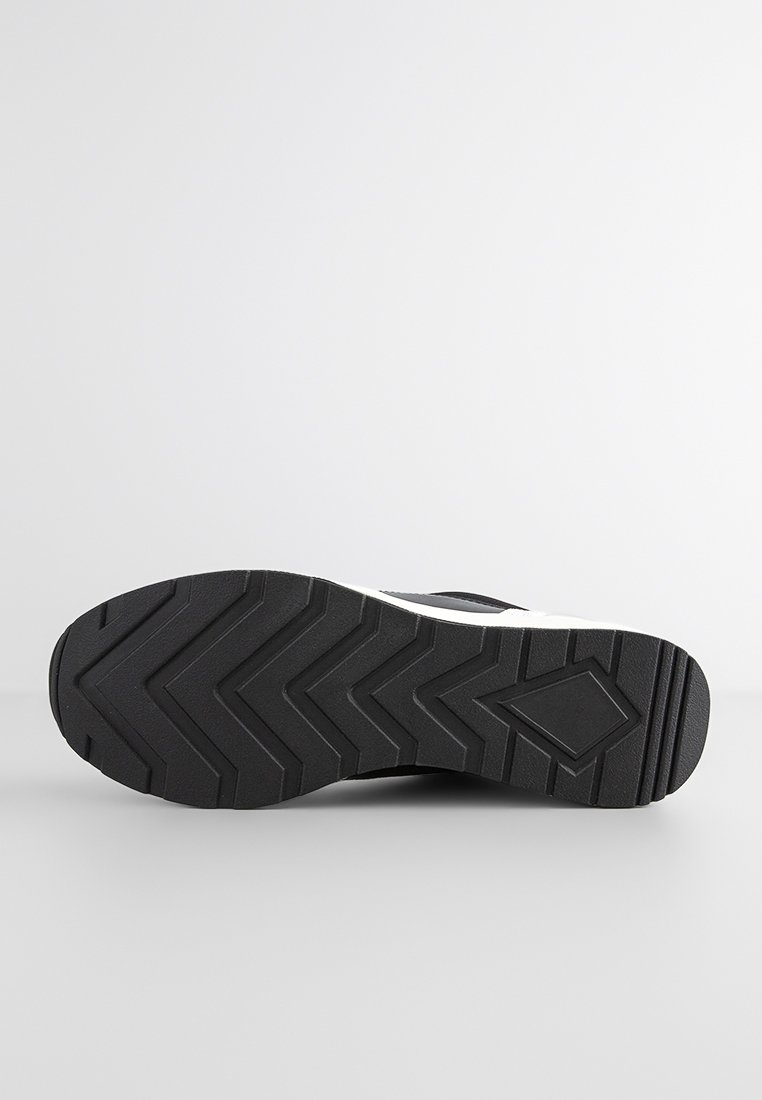 Semelle noire de sneaker avec une gomme texturée, présentant un motif de bande de roulement en zigzag et un accent blanc près du bord supérieur.