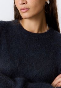 Pull en maille bleu foncé, doux et pelucheux, avec un col rond. Présente une texture subtile et un design simple et épuré. Des accents de bijoux en argent visibles.