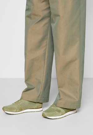 Ben iført løse, beige bukser og lysegrønne mesh- og ruskindssneakers med hvide såler på en ensfarvet grå gulv.