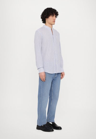 Srajca z dolgimi rokavi, prugasta v beli in modri barvi, z mandarin ovratnikom, v kombinaciji s svetlo modrimi kavbojkami in črnimi čevlji.