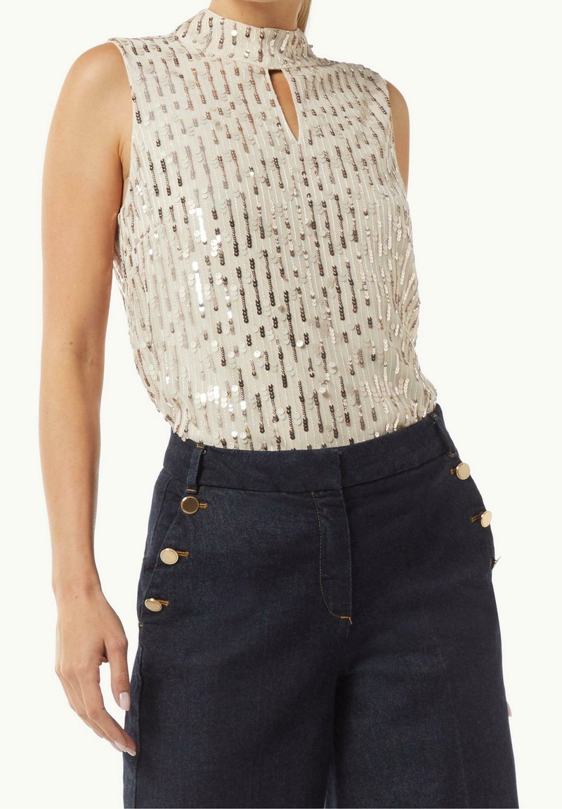 Haut blanc sans manches avec sequins argentés verticaux et petit décolleté en trou de serrure, associé à un pantalon taille haute foncé avec des boutons dorés.