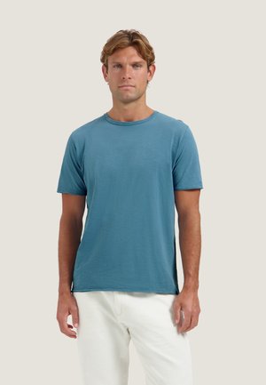 Uomo con capelli castano chiaro che indossa una maglietta blu a maniche corte e pantaloni bianchi, in piedi davanti a uno sfondo chiaro e semplice.
