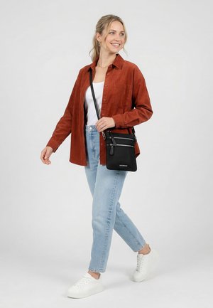 Lächelnde Frau geht, trägt ein rostfarbenes Hemd, weißes Oberteil, helle Jeans, weiße Turnschuhe und eine schwarze Umhängetasche mit silbernen Reißverschlüssen vor einem einfarbigen Hintergrund.