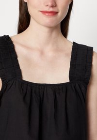 Top preto sem mangas com decote quadrado, tecido franzido com um design de alças texturizadas e com babados. Material leve, caindo suavemente.