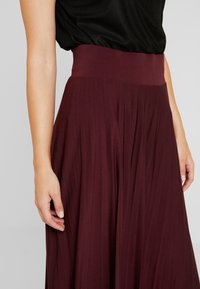Donna che indossa una gonna burgundy a vita alta e fluida, con una maglietta nera infilata dentro, le mani appoggiate ai fianchi su uno sfondo grigio.