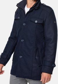 Man wearing a navy blue buttoned pea coat with flap pockets, epaulets, and a zip-up collar over a white shirt and grey pants.